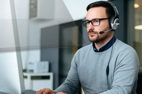 A professional man wearing a headset and glasses, working at a computer in a modern office environment, focused on providing tech support.