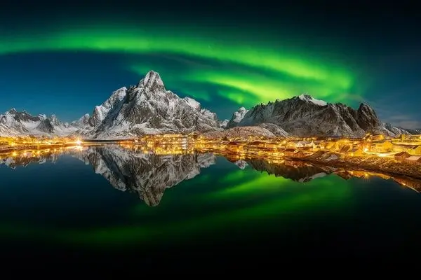 A wooden cabin sits in a snowy field under a vibrant green and purple aurora borealis spanning the night sky.