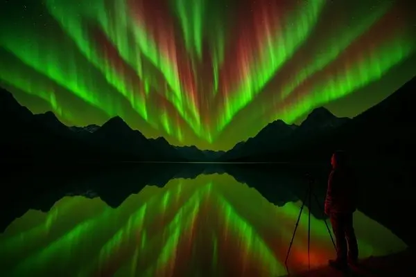 where to see the northern lights: A person stands by a camera tripod, gazing up at spectacular vertical green and red aurora borealis reflected in a calm lake.
