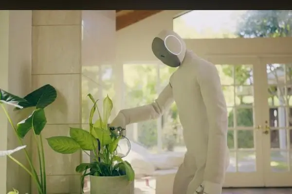 A humanoid robot (the same model as in Image 4, identifiable by the pale, cloth-like body) performing a simple household task indoors.