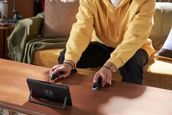 Man in yellow hoodie playing Nintendo Switch 2 in tabletop mode on wooden table with detached Joy-Con 2 controllers.
