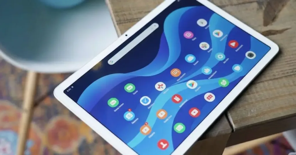 A Google Pixel Tablet with a white bezel lying on a light wood surface. The tablet screen shows a bright blue abstract wave wallpaper with a dense grid of colorful Android app icons and a Google search bar at the top. The background is softly blurred, showing a white chair leg and a blue decorative object. A Droid Life watermark is in the lower right corner.