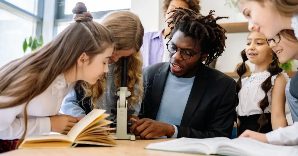 artificial intelligence basics: : A teacher helping a group of elementary school students examine a book and a microscope during a classroom science activity.