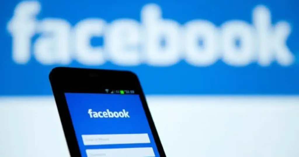meta ai A smartphone displaying the Facebook login screen held in front of a blurred computer monitor showing the Facebook logo.