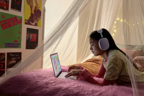 A young student using a pink MacBook Neo while lying on a bed and wearing large white headphones.