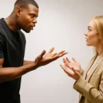 how to stay calm: A Black man in a black t-shirt passionately speaks with open hands to a blonde woman in a beige blazer who looks back at him with a surprised or defensive expression.